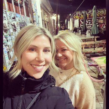 Honey Elixir Bar - Two women with blonde hair are smiling and taking a selfie outside at night, with a background of outdoor seating, umbrellas, and graffiti. - Lounges Near Me - Denver, Colorado