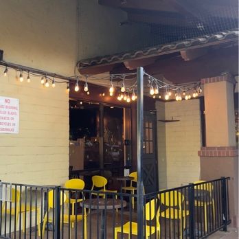 Paste Thai - Patio seating - A small outdoor patio features yellow chairs, a table, string lights, and a black metal fence, adjacent to a building with a glass door. - Thai Near Me - Davis, California