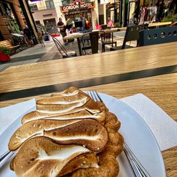 Lemon Meringue - A plate of toasted bread with marshmallow topping is placed on a wooden table in an outdoor seating area of a coffee shop. The background features a lively street scene with people, tables, and chairs, along with signs for businesses like Gordon Hotel and Magpie Coffee Shop. There are musicians performing live music, and the area is decorated with potted plants and storefronts, creating a vibrant atmosphere.