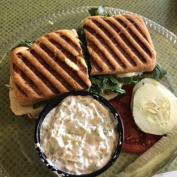 Culinary Fox Cafe - Veggie Grinder - A sandwich features two slices of grilled bread with visible grill marks, filled with fresh lettuce, tomato, and cucumber slices. Accompanying the sandwich is a small black container of creamy herb dip, and the plate also contains additional cucumber and tomato slices. The food is served on a clear plastic plate placed on a green textured surface. - Cafes Near Me - Elgin, Illinois