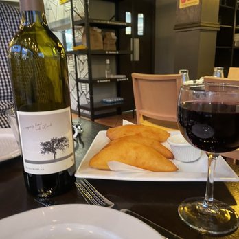 Bienmesabe - The image shows a dining table with a bottle of wine, a glass of red wine, and a white rectangular plate holding three empanadas with a small container of dipping sauce. The background includes chairs, a shelf, and a wall with posters, indicating a restaurant environment. - Venezuelan Near Me - Chicago, Illinois
