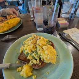A plate of scrambled eggs served on a slice of toasted bread, garnished with black pepper, is placed on a dark wooden table in a restaurant. The background shows other diners, drinks, and a bright, modern interior with white columns and warm lighting. The table also has glasses of water, a berry drink, and condiments, indicating a casual brunch setting.