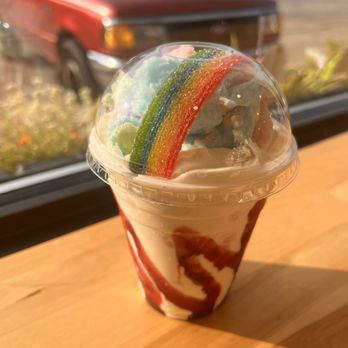 Better Together Cafe - A milkshake in a clear plastic cup features a swirl of vanilla and caramel syrup, topped with rainbow gummy candies and colorful sprinkles. The drink is set on a wooden surface near a window, with a blurred background showing a red vehicle outside. The toppings include a rainbow gummy candy and pastel-colored cotton candy, creating a vibrant presentation. - Desserts Near Me - South Milwaukee, Wisconsin