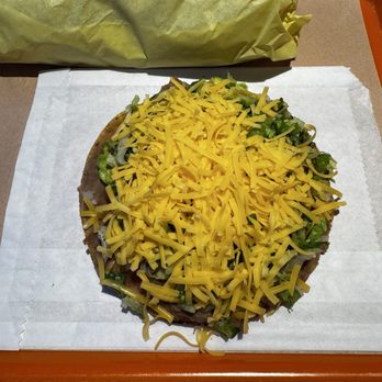 Darlene's Veg - Secret menu Tostada!!! So good and satisfying if you're nostalgic for this! Vegan or real cheese! - A close-up of a tostada topped with shredded yellow cheese, chopped lettuce, and other ingredients, placed on a paper sheet on an orange tray. In the background, there is a wrapped item, possibly a burrito or sandwich, on a tray. The setting appears to be a casual dining environment, likely a diner or fast-food restaurant. - Vegetarian Near Me - Eugene, Oregon