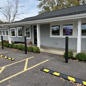 Half Baked Cafe - Front of cafe - The image shows the exterior of a single-story commercial building with gray siding and white trim. There are multiple windows, one of which displays a neon 'Open' sign. The entrance is centered, with pumpkins placed nearby, suggesting a seasonal decoration. The parking lot in front has marked parking spaces and black and yellow wheel stops, with some fallen leaves scattered on the asphalt. Trees with sparse foliage are visible in the background, indicating an autumn setting. - Ice Cream & Frozen Yogurt Near Me - Lockport, New York