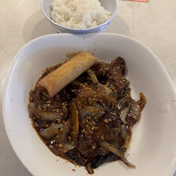 Upside on Moore - A meal consisting of beef with onions and a spring roll on a white plate, with a bowl of white rice placed behind the plate. The dish appears to be a stir-fry with a dark sauce and chopped nuts or garlic on top. The rice is served in a small bowl, and a business card or ticket is visible in the background. - Food Court Near Me - Arlington, Virginia