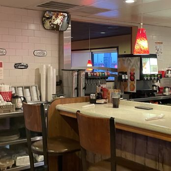 Denny's - The image shows the interior of a coffee shop or diner with a curved counter and wooden chairs. Behind the counter, there are beverage dispensers, a small mirror on the wall, and various supplies such as cups and condiments. The decor includes red pendant lights and a window in the background, creating a casual dining atmosphere. - American Near Me - Raton, New Mexico