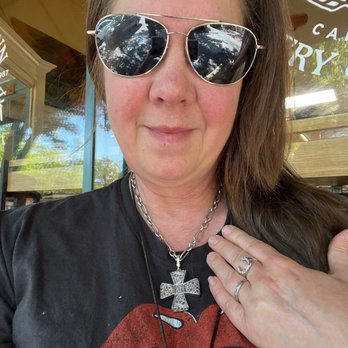 Favorites Consignment - My new cross!!!!!!! - A woman with long brown hair, wearing aviator sunglasses, a silver cross necklace, and rings, sitting outside a cafe. - Used, Vintage & Consignment Near Me - Healdsburg, California