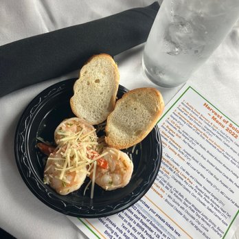 Bugeye Grill - Pesto shrimp sautee - A black plastic plate contains two slices of bread and two shrimp topped with shredded cheese and garnished with small pieces of tomato. The plate is on a white tablecloth, accompanied by a tall glass of ice water and a printed menu with colorful text. A black napkin is also visible nearby. - Seafood Near Me - Solomons, Maryland