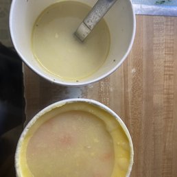 Top - Olympus Grill Bottom - another local place my sister loves - The image shows two cups of soup on a wooden table. The top cup contains a light-colored, creamy soup with a metal spoon inserted. The bottom cup holds a different soup, possibly a broth or consomme, with a thin, translucent surface. The table surface is wooden, and there is a plastic or glass surface in the background. The photo is taken from above, capturing the cups and part of the table.