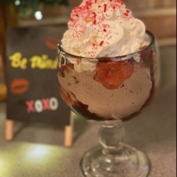 Sweeties - A dessert in a clear glass cup features whipped cream topped with red sprinkles and strawberries inside. The background includes a blurred chalkboard sign with colorful writing and a floral arrangement, indicating a cozy restaurant or cafe setting. The dessert appears to be a layered treat, possibly ice cream or pudding, garnished with whipped cream and fruit. - Desserts Near Me - Corbin, Kentucky