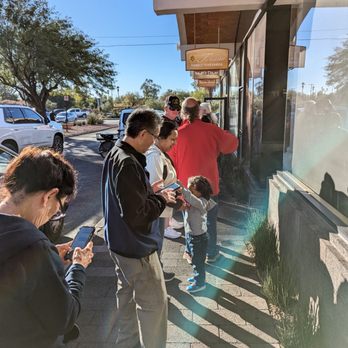 Barrio Bread - Friday Morning Line - A group of people, including adults and children, are waiting in line outside a storefront on a sunny day, with some using their phones. - Bakeries Near Me - Tucson, Arizona