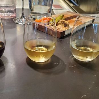 Brooklyn Winery - Three wine glasses with red and white wine are placed on a dark table, with food and drink items visible in the background. - Venues & Event Spaces Near Me - Brooklyn, New York
