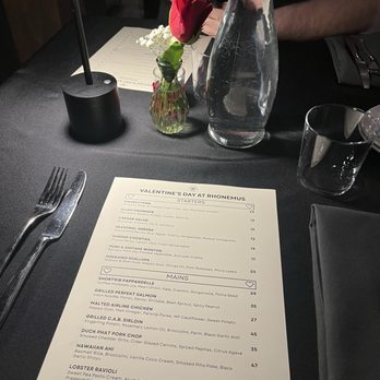Rhonemus Cellars - A table at Rhonemus Cellars displays a printed menu, a small vase with a red flower and white filler, a glass water bottle, and a drinking glass. A person with a dark shirt and arm is partially visible in the background, and a black table lamp is also present. The setting suggests a dining experience in a winery environment with a focus on wine and food offerings. - Wineries Near Me - Lancaster, Ohio