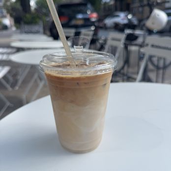 Coffee Check - Iced latte - A clear plastic cup contains iced latte with visible layers of coffee and milk, topped with ice cubes and a straw. The cup is on a white round table outdoors, with empty chairs and trees in the background, suggesting a café patio setting. - Coffee & Tea Near Me - Brooklyn, New York