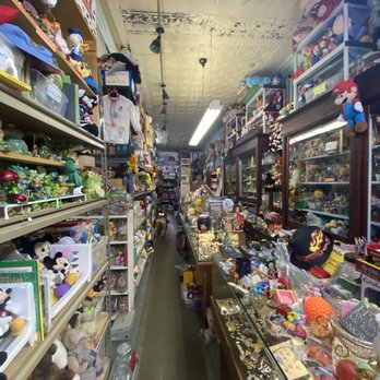 Aldridge's - A narrow aisle in a thrift store or antique shop is densely packed with toys, figurines, and collectibles. Shelves on the left are filled with plush toys, Disney characters, and small decorative items, while display cases on the right contain jewelry, small trinkets, and vintage memorabilia. The ceiling shows signs of wear with peeling paint and an old tin ceiling design. Fluorescent lighting illuminates the cluttered space, creating a busy and nostalgic atmosphere. - Antiques Near Me - Tuscola, Illinois