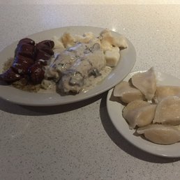 Cabbage Rolls - Two plates of Polish cuisine are shown on a white textured table. The larger plate contains meat, possibly kielbasa, with a dark sauce, along with potato dumplings and a creamy mushroom sauce with mushrooms. The smaller plate holds several pierogi, a type of Polish dumpling, with a light, slightly shiny surface. The presentation is simple, with no additional garnishes or decorations.