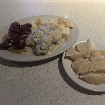 Szarotka - Cabbage rolls with Mushroom sauce, kielbasa and kraut, Polish Dumplings, and Sweet Cheese Pierogos - Two plates of Polish cuisine are shown on a white textured table. The larger plate contains meat, possibly kielbasa, with a dark sauce, along with potato dumplings and a creamy mushroom sauce with mushrooms. The smaller plate holds several pierogi, a type of Polish dumpling, with a light, slightly shiny surface. The presentation is simple, with no additional garnishes or decorations. - Bars Near Me - Justice, Illinois