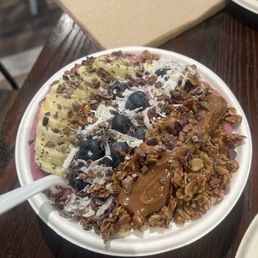 Acai Bowl - A smoothie bowl with a pink-colored base is topped with sliced bananas, blueberries, granola, shredded coconut, chocolate shavings, and a dollop of peanut butter. The bowl is placed on a dark wooden table, with a white spoon inside it. In the background, there is a closed napkin holder and part of another plate.