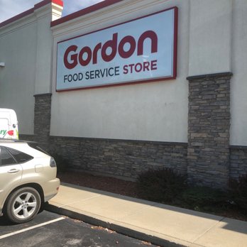 GFS Marketplace - Welcome - The image shows a large rectangular sign for Gordon Food Service Store mounted on the exterior wall of a beige building. The sign features the store's name in bold red and black letters, with additional text indicating it is a food service store. The building has decorative stone accents at the lower part of the wall. In front of the building, there are parking spaces with cars parked, and a sidewalk runs along the front of the store. The sky above is partly cloudy. - Grocery Near Me - Darien, Illinois