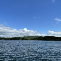 The lake view - A landscape featuring a large body of water with gentle ripples, bordered by green hills in the background. The sky above is partly cloudy with patches of blue visible. The scene captures a peaceful outdoor setting, likely near a natural or recreational area.