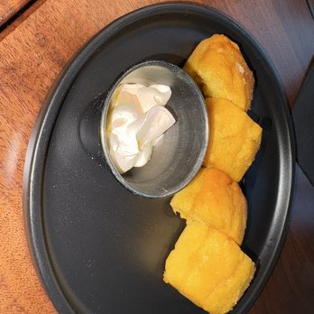 Shiso - Cornbread - Three pieces of cornbread are arranged on a black oval plate, accompanied by a small metal cup of whipped cream or butter, on a wooden table. - Cocktail Bars Near Me - Miami, Florida
