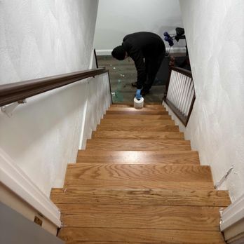 Modern Pest Services - A person dressed in black clothing, including a hoodie and mask, is bent over at the bottom of a wooden staircase, cleaning with a roll of toilet paper. The staircase has wooden steps and white walls, with a handrail on the left side. The area at the bottom of the stairs appears to be a hallway or room with some clutter and a bicycle in the background. - Pest Control Near Me - Woburn, Massachusetts