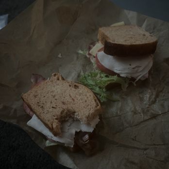Panera Bread - Turkey bacon bravo - A sandwich featuring turkey bacon, lettuce, tomato, and cheese on whole wheat bread is shown, with a bite taken out of one half. The sandwich is placed on crumpled brown paper, and the setting appears to be in a dimly lit environment, possibly near a vehicle or outdoor area. - Sandwiches Near Me - Peoria, Illinois