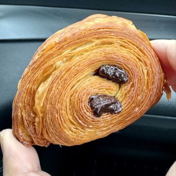 Tangy Cafe - Pain au chocolat - A flaky, golden-brown pain au chocolat with two visible chocolate pieces inside, held by a person's hand against a dark background. - Cafes Near Me - Markham, Ontario
