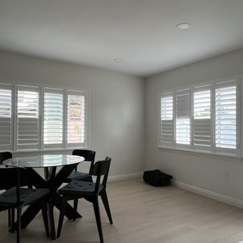 Sun Blinds - A bright dining space features a round glass table with four black chairs, situated near two windows with white plantation shutters, in a room with white walls and light-colored flooring. - Shades & Blinds Near Me - Baldwin Park, California