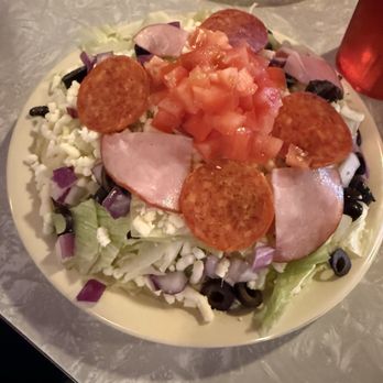 Pizza House - Deluxe Salad - A salad on a plate includes chopped lettuce, diced tomatoes, slices of ham, pepperoni, shredded cheese, and black olives. The ingredients are arranged with toppings in the center and leafy greens around the edges. The salad appears fresh and is served on a light-colored plate, with a textured table surface visible underneath. - Pizza Near Me - Springfield, Missouri