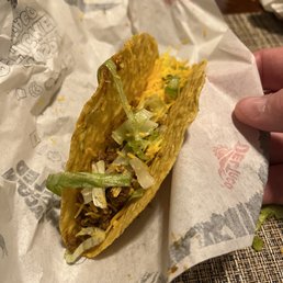 Comedy tacos - A taco with a hard taco shell contains shredded lettuce, shredded cheese, and seasoned ground beef. The taco is held by a person's hand and rests on branded paper, with some crumbs and ingredients visible around. The background includes a woven placemat and a wooden surface.
