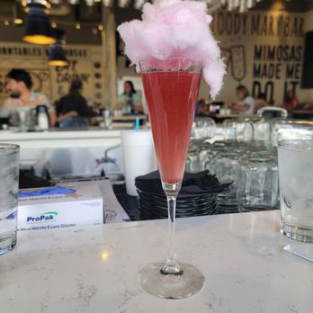 Hash Kitchen - Blackberry mimosa cotton candy glitter bomb - A pink-colored cocktail with cotton candy on top is presented in a tall, narrow glass. The drink appears fizzy or sparkling, and the cotton candy is fluffy and pink, adding a decorative element. The setting is a bar or restaurant with a busy atmosphere, visible patrons, and a modern interior design with hanging lights and wall art. - Breakfast & Brunch Near Me - Chandler, Arizona