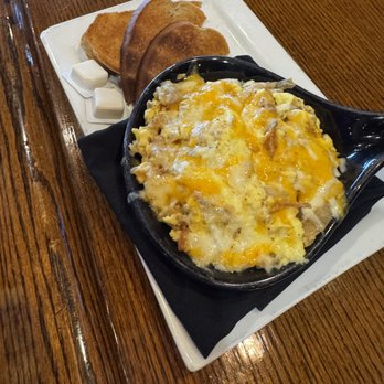 Stewart's Brewing - Breakfast Skillet - A breakfast meal presented in a black skillet containing scrambled eggs topped with melted cheese, possibly including meat or vegetables. The skillet is placed on a white rectangular plate with a black napkin underneath. Alongside, there are three slices of toasted bread with butter pats on a separate white plate. The setting appears to be in a restaurant or pub environment with a wooden table. - Brewpubs Near Me - Bear, Delaware