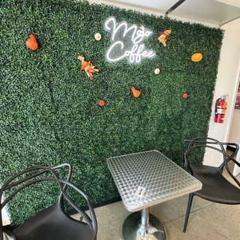 Mojo Coffee - Inside - A green artificial hedge wall serves as a backdrop, decorated with a neon sign that reads 'Mojo Coffee' and various fall-themed ornaments such as small pumpkins and decorative figures. In front of the wall, there are two black chairs with curved armrests and a small metallic table with a textured surface. The setting appears to be an indoor seating area within a coffee shop or cafe. - Coffee & Tea Near Me - Round Rock, Texas