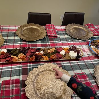 Mila's Mesa - Charcuterie board - A rectangular dining table is covered with a red, green, and white plaid tablecloth. It is set with multiple gold-colored plates, each placed in front of a chair. In the center, there is a long platter with assorted cheeses, grapes, and other snacks. To the right, there is a bowl with olives, cheese, and other items. A child's hand, wearing a dark sleeve with a floral pattern, is pointing at one of the plates. Two black chairs are visible at the head of the table, and a person is partially visible standing to the right. - Caterers Near Me - Albuquerque, New Mexico