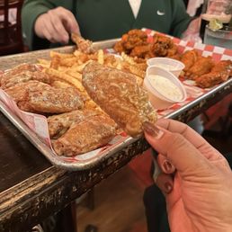 The yummiest chicken wings! - A person is holding a fried chicken wing over a tray filled with various fried chicken pieces, French fries, and dipping sauces. The tray is lined with checkered paper, and the background shows a busy restaurant with other diners and drinks on the table. The focus is on the food and the hand holding the wing, with a person in a green top visible in the background.