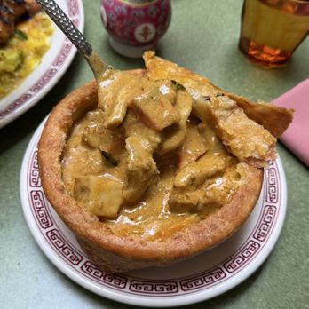 Kelang - Curry Chicken Pot pie - A bread bowl filled with curry chicken stew, featuring chunks of chicken and vegetables in a thick, creamy sauce. The bread bowl is placed on a decorative plate with a red geometric border, on a green table. In the background, there is a small vase with flowers, a glass of iced tea, and other dishes, indicating a restaurant setting. - Salad Near Me - Brooklyn, New York