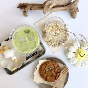 Studio De Corner - The image shows a white table with various items including a chocolate chip cookie with googly eyes on a paper napkin, a glass of iced green tea with ice cubes, dried flowers in a glass, and plush toys resembling a sheep and a smiling flower. A piece of driftwood is also present on the table, along with a small plate and a tray. The setting appears decorative and playful, with a focus on aesthetic arrangement. - Cafes Near Me - Markham, Ontario