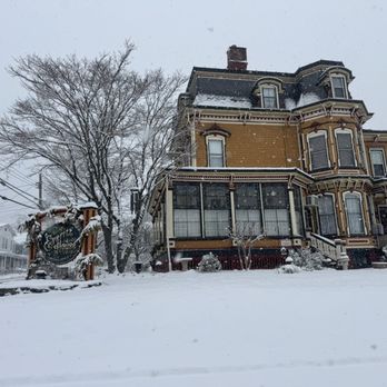 Everitt House - Outside on a snowy morning - A Victorian-style house built in the Second Empire architectural style features decorative trim and multiple bay windows. The house is painted in a yellowish color with contrasting trim, and it is covered in snow. Snow is falling, and the yard, trees, and surrounding area are blanketed in snow, indicating a winter scene. - Bed & Breakfast Near Me - Hackettstown, New Jersey