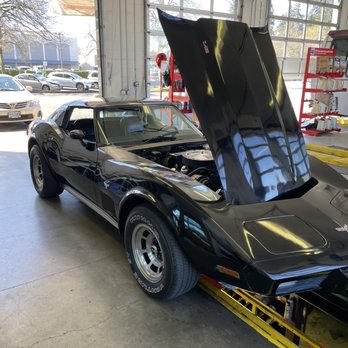 First Lube Plus - Classic Corvette oil change - A black Chevrolet Corvette with its hood raised is inside an automotive service garage, with the engine visible and the garage door open to the outside. - Oil Change Stations Near Me - Salem, Oregon