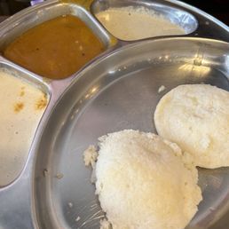 Idly - Two soft, round idlis are positioned on a stainless steel plate, with three small compartments containing different chutneys and sambar. The chutneys include a white coconut chutney, a creamy white or beige chutney, and a darker, spicy sambar. The image captures a typical South Indian breakfast presentation, emphasizing the variety of flavors served with idlis.