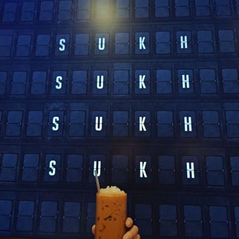 Sukh - Vibe - A person holds a cup of bubble tea in front of a wall with the word 'SUKH' displayed multiple times. - Thai Near Me - Brooklyn, New York