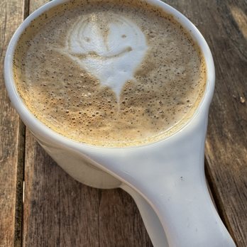 High/Low - A latte with foam art depicting a smiley face is in a white ceramic mug placed on a wooden table. The foam art is created with steamed milk and coffee, with a smiley face design on the surface. A glass of water is positioned behind the mug, partially visible in the background. The setting appears to be outdoors or in a rustic environment, with natural lighting and a textured wooden surface. - Breakfast & Brunch Near Me - Oceanside, California