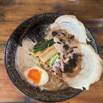 Tsuruya - Black garlic Tonkatsu - A bowl of ramen with a creamy broth contains sliced pork, a halved soft-boiled egg with a runny yolk, green vegetables, bamboo shoots, and black garlic oil. The bowl has a decorative black and gold pattern, and the setting includes a wooden table with a glass of water in the background. - Ramen Near Me - Berkeley, California