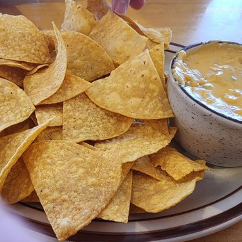 Christy Mae's Restaurant - Chips and queso - A large plate holds numerous tortilla chips, with some being broken or folded, and a bowl of melted cheese dip placed beside the chips. The setting appears to be a casual dining environment with a wooden table surface and some paper napkins or packaging in the background. - Sandwiches Near Me - Albuquerque, New Mexico
