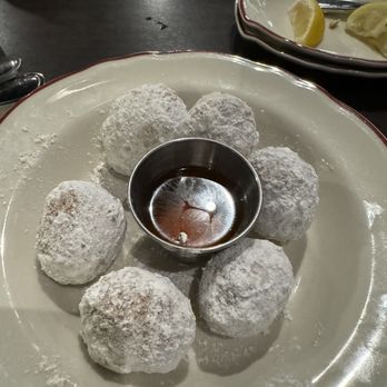 Tony C's - A round ceramic plate holds six powdered sugar-coated Italian dessert balls arranged in a circle around a small metal container of dark dipping sauce. The plate is on a dark wooden table, with a smaller dish containing lemon wedges visible in the background. The setting suggests a restaurant serving Italian or American-Italian cuisine. - Pizza Near Me - Austin, Texas