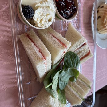 Lovey's Tea Shoppe - Strawberry jam sandwich and cucumber dill with some jam and cream. The scones were so soft!! Who knew!! - A rectangular clear plastic tray contains an assortment of finger sandwiches with white bread and jam filling, garnished with fresh greens. Two small bowls hold clotted cream with a blackberry and jam with a strawberry. The tray is placed on a pink tablecloth, and part of a cup with a pastry is visible on the side. The presentation suggests a traditional British-style tea service. - British Near Me - Pacifica, California