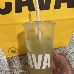Cucumber Mint Lime Juice - A person is holding a clear plastic cup containing cucumber mint lime juice, with visible ice and slices of cucumber inside. The cup has a white straw and the word 'CAVA' printed on it. The background features a yellow shopping bag with the same 'CAVA' branding, placed on a speckled granite countertop. The person's hand is partially visible, wearing a pink sleeve.