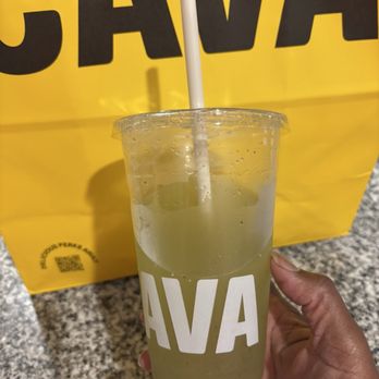 CAVA - Cucumber Mint Lime Juice - A person is holding a clear plastic cup containing cucumber mint lime juice, with visible ice and slices of cucumber inside. The cup has a white straw and the word 'CAVA' printed on it. The background features a yellow shopping bag with the same 'CAVA' branding, placed on a speckled granite countertop. The person's hand is partially visible, wearing a pink sleeve. - Mediterranean Near Me - Arlington, Texas